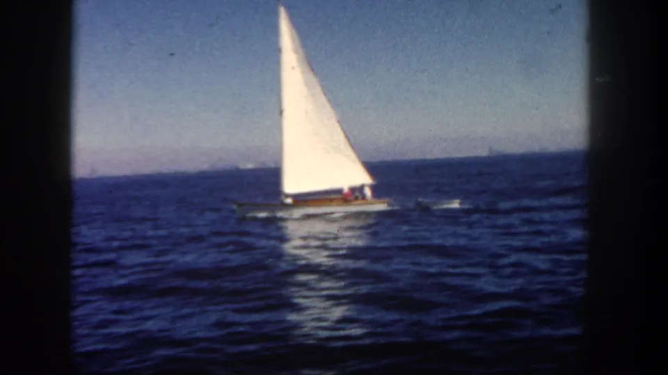 The 1962 San Diego home-movie frame centers a small sailboat on open water with a hazy horizon and dark film edge.