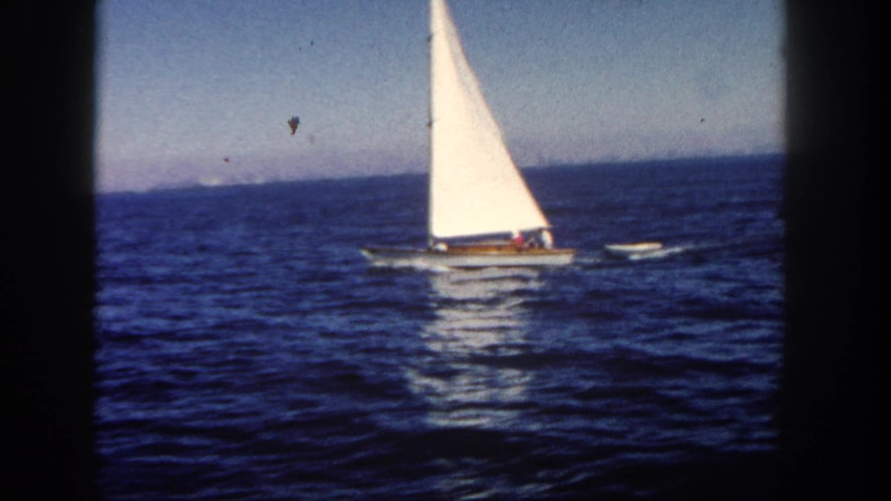 1962 San Diego 8mm frame of a sailboat on open blue water with a small dinghy trailing behind it.