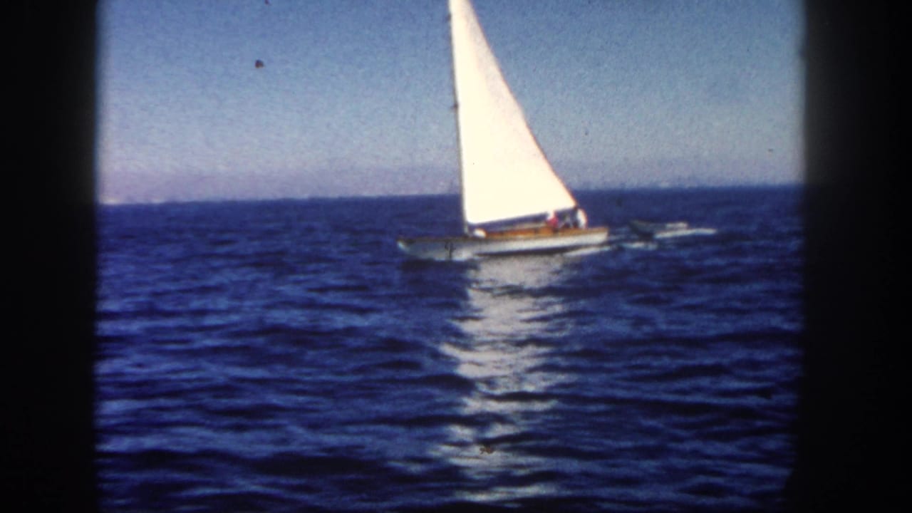 1962 San Diego home-movie frame showing a sailboat under clear sky, towing a dinghy across open water.