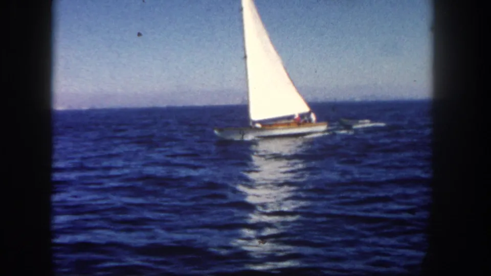 1962 San Diego home-movie frame showing a sailboat under clear sky, towing a dinghy across open water.