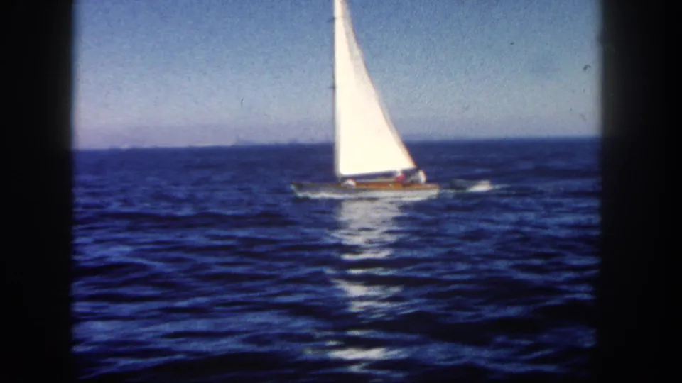 1962 San Diego 8mm still of a small sailboat riding across choppy water with a dinghy in tow.