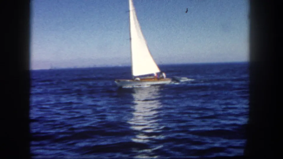 1962 San Diego coastal home-movie frame of a sailboat and trailing dinghy on open blue water.