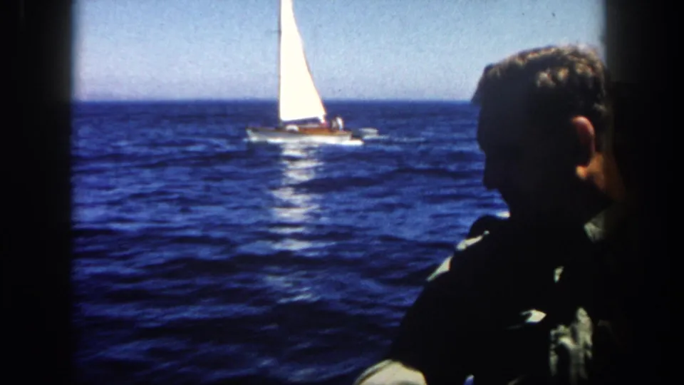 1962 San Diego frame with a white sailboat on blue ocean and an adult man seen in shadowed profile from a nearby vessel.