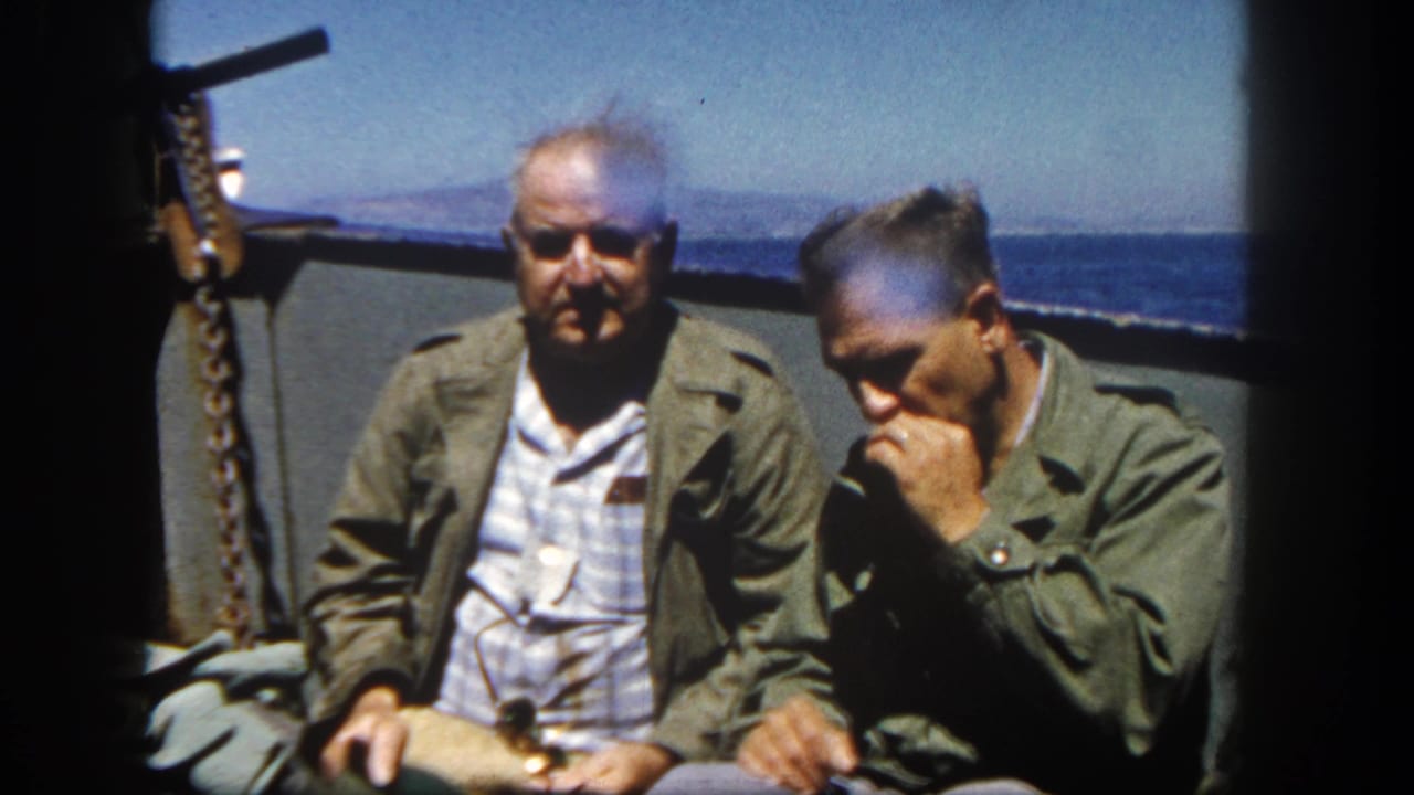 In 1962 San Diego-area 8mm footage, two adult men in olive jackets sit on a ship deck against open water and distant land.