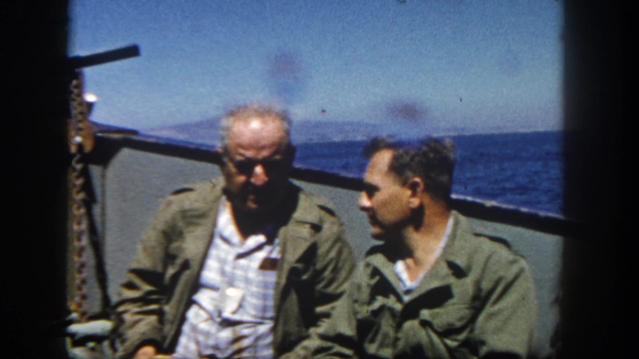 Two adult men sit close together on a ship deck near San Diego, one facing the other as blue water and distant shoreline fill the background.