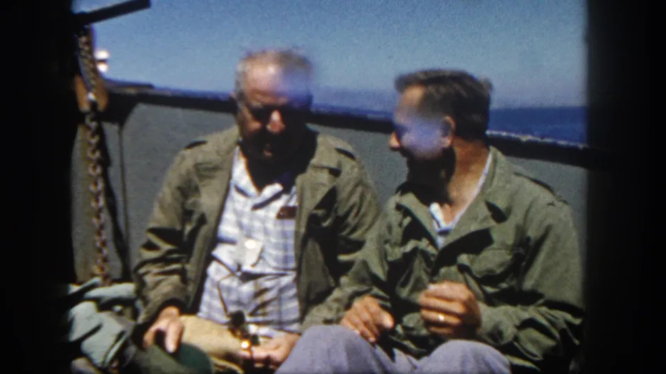 The older man looks down while the younger man leans toward him on a sunlit ship deck, with sea and railing visible in the 8mm frame.