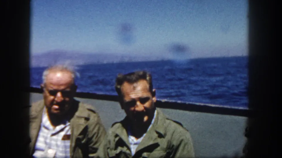 Two men in olive outerwear face the camera on a ship deck, with open blue sea and low distant hills visible in this 1962 San Diego home-movie frame.
