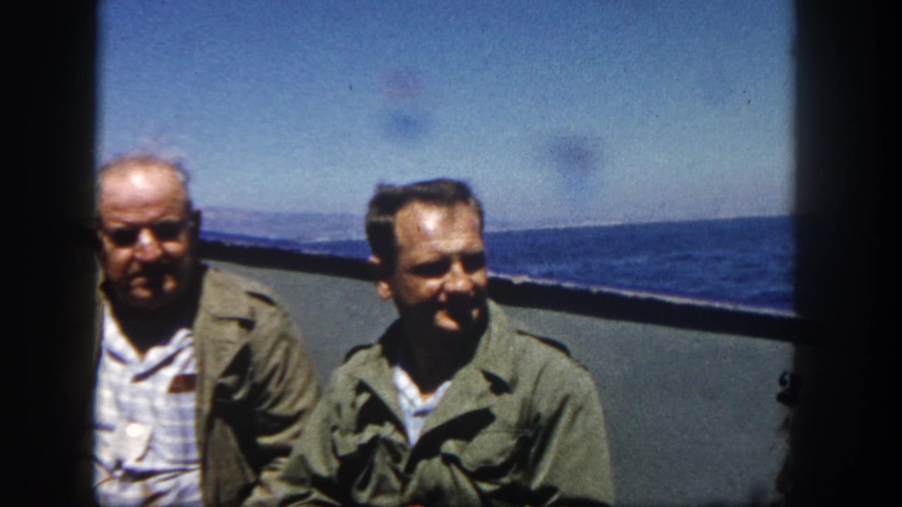Two adult men sit or stand near a boat rail with ocean and distant coast behind them.