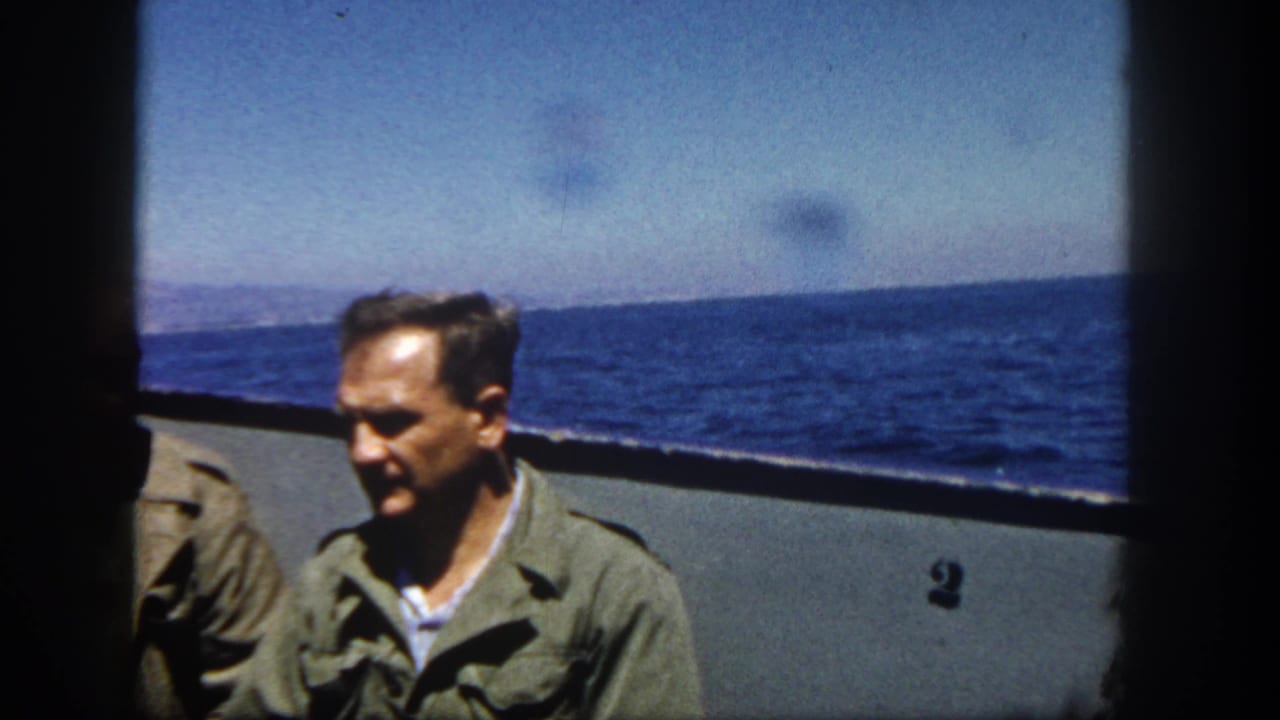 A man in a green jacket sits near a boat rail with open sea behind him.