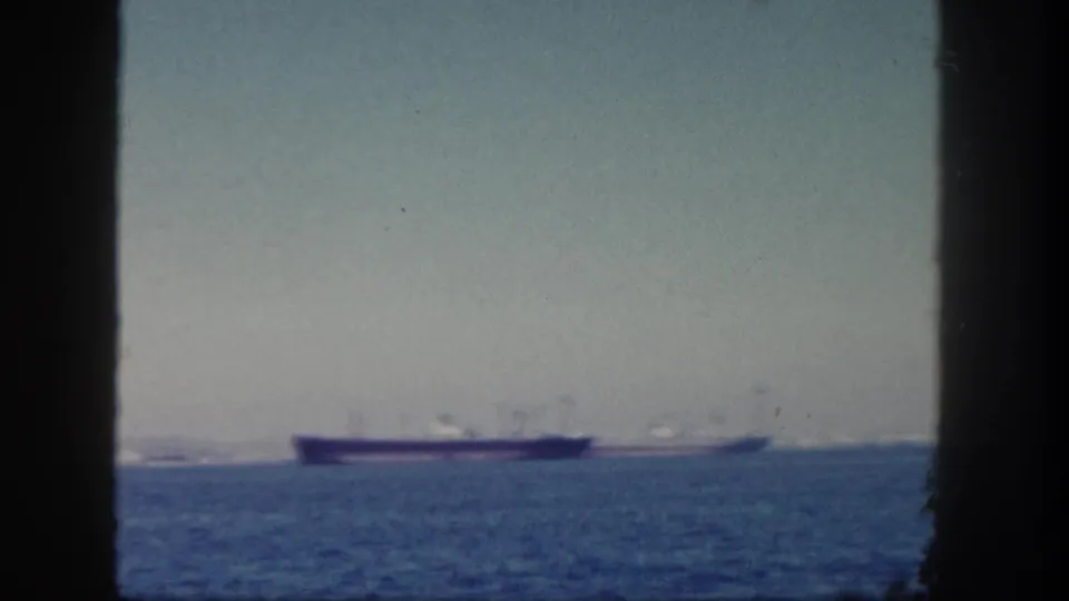 Distant cargo ship on the water in 1962 San Diego-area 8mm footage, framed by dark ship edges.