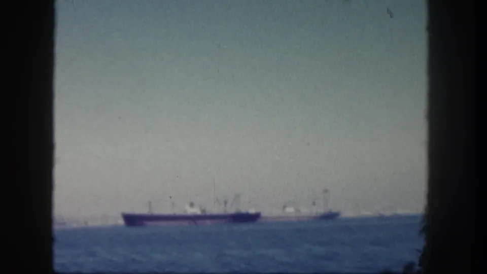 1962 San Diego 8mm frame with a large cargo ship and smaller vessel near the horizon on a calm ocean.