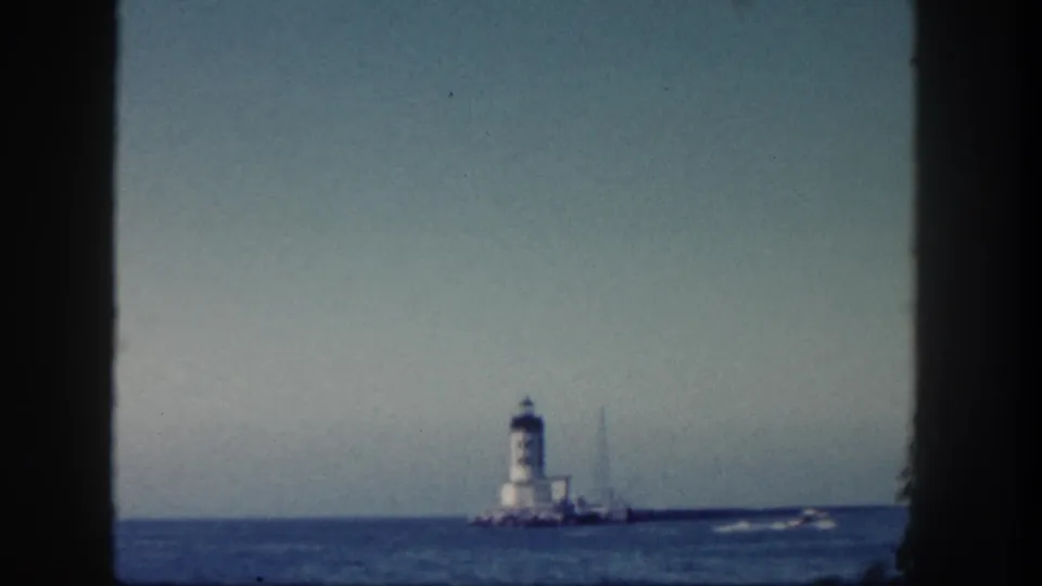 1962 San Diego 8mm frame showing a distant lighthouse and small boat from a vessel-window viewpoint.