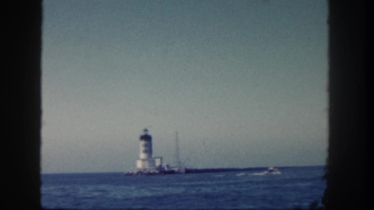 Distant white lighthouse on a rocky jetty with ocean water and a pale sky, framed by dark film borders.