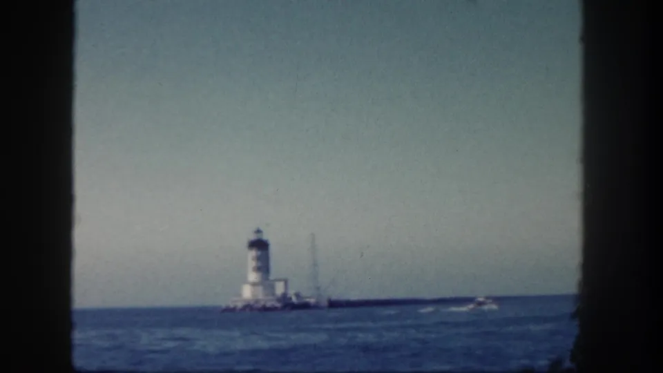 1962 San Diego coastal home-movie frame showing a distant white lighthouse, rocky jetty, blue ocean, and pale sky through dark frame edges.