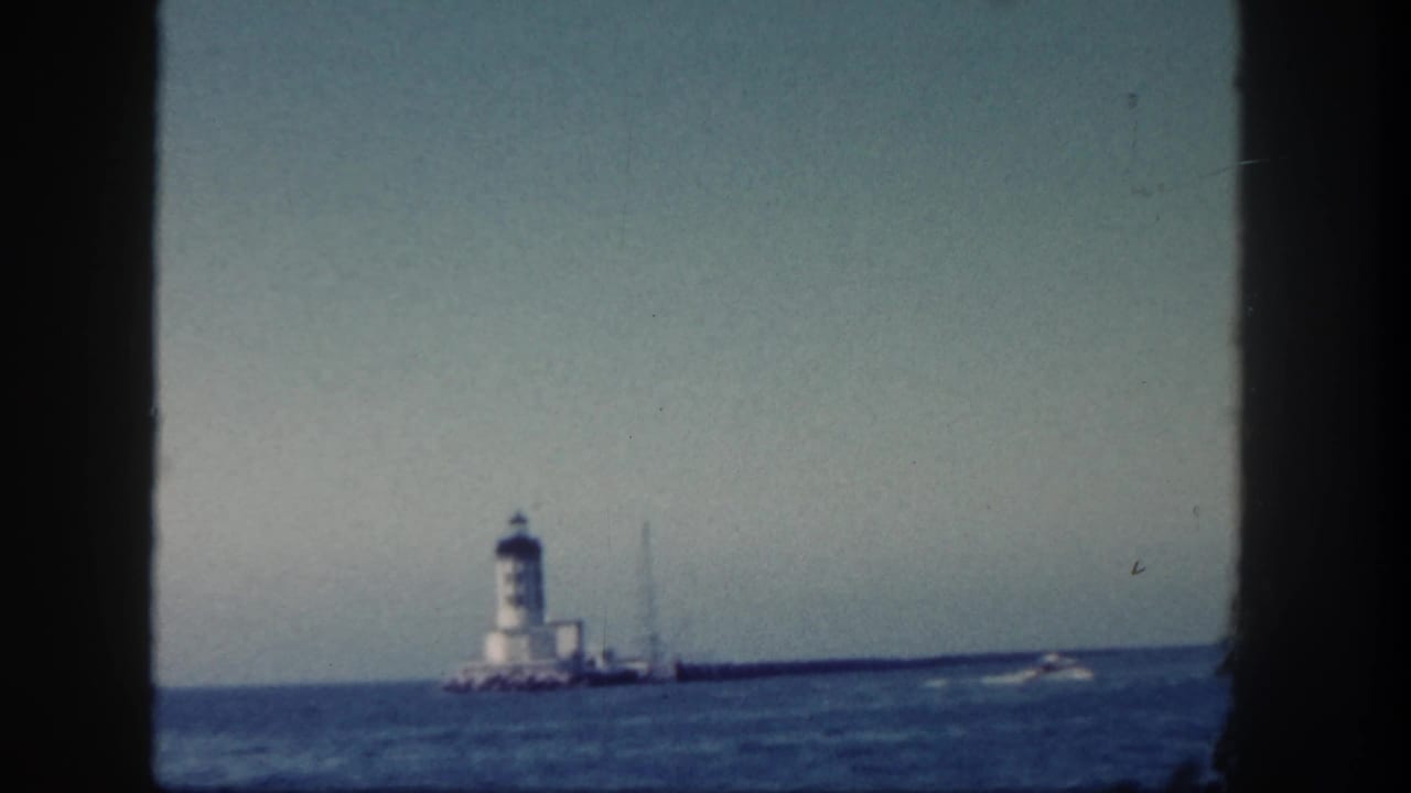 White lighthouse and jetty in the distance with a small boat wake crossing the ocean to the right.