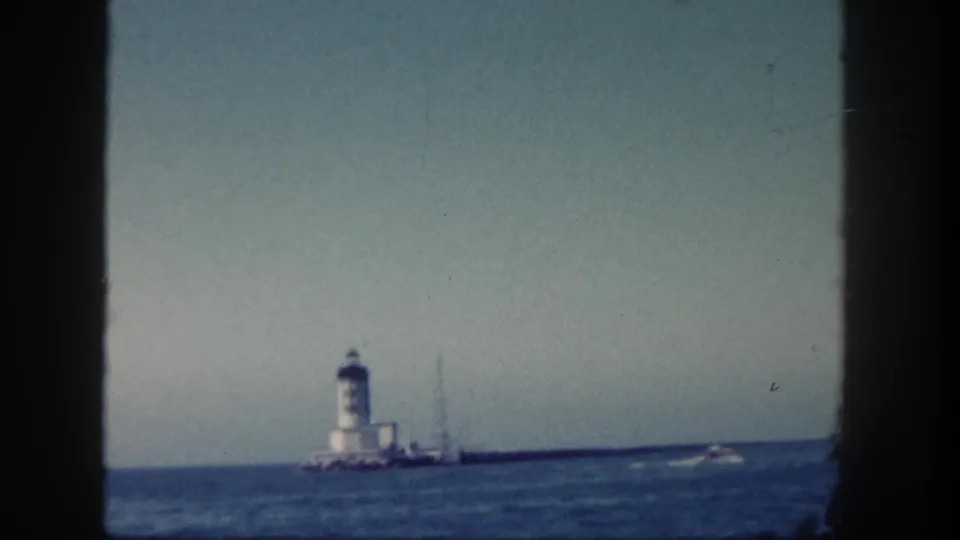 1962 San Diego coastal view with a lighthouse on a jetty and a small speedboat wake moving across the water at right.