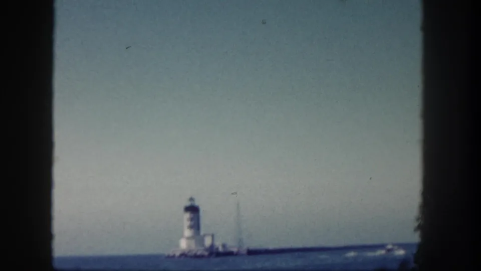 1962 San Diego 8mm frame showing a distant lighthouse and breakwater across calm blue ocean under a clear pale sky.