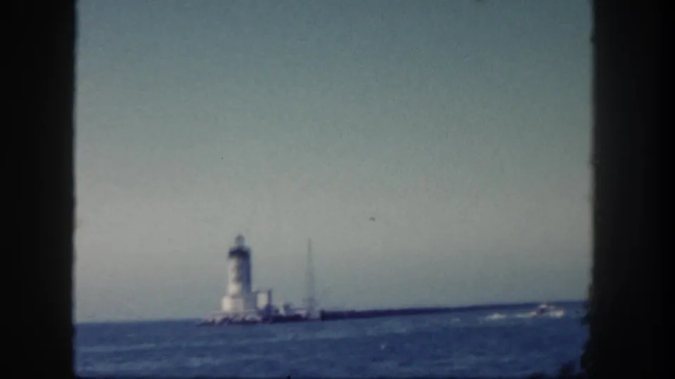 1962 San Diego coastal frame with lighthouse, long jetty, ocean horizon, and a small boat or wake at the right edge.