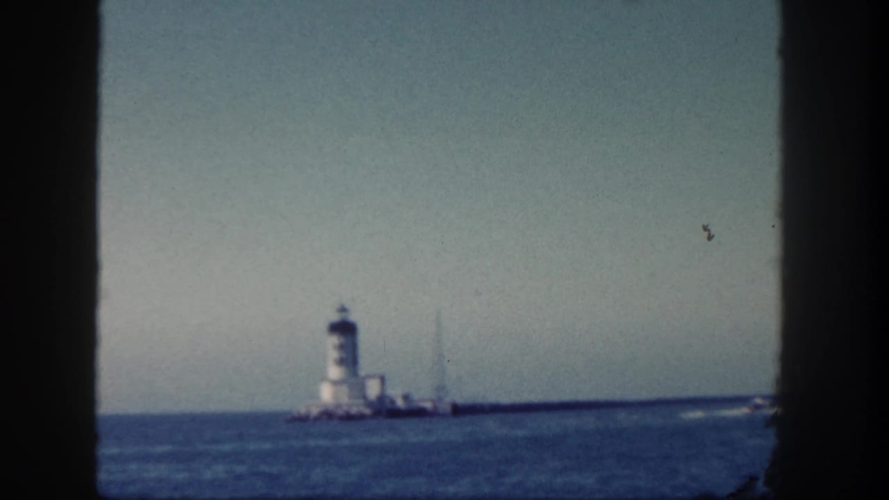 Distant white tower on a rocky pier across blue water, framed by dark vertical edges.