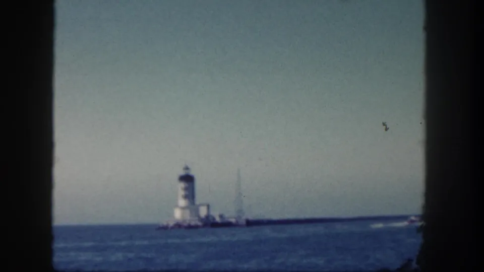 1962 San Diego home-movie frame showing a distant white lighthouse-like tower on a breakwater across calm blue water.