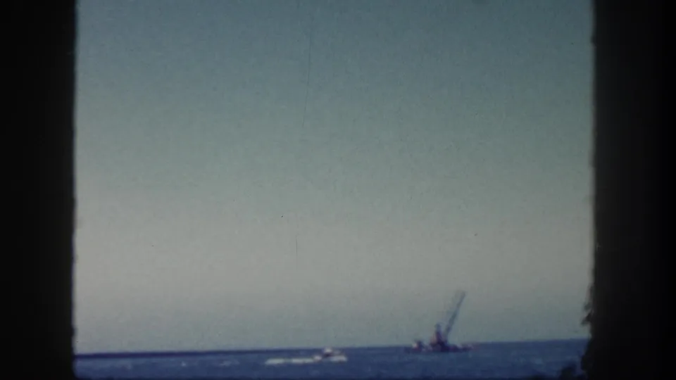 1962 San Diego coastal frame with open water, a small boat wake, and a distant crane-like silhouette near the horizon.