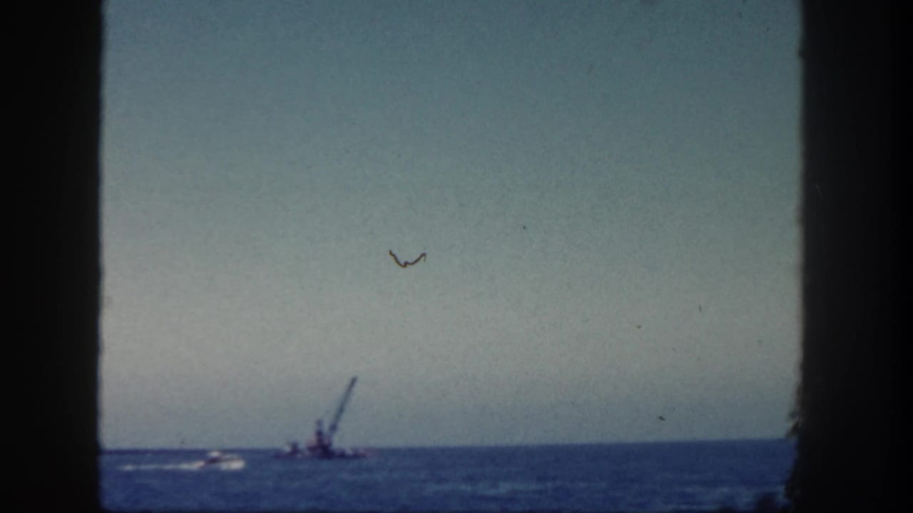 1962 San Diego 8mm frame showing a distant crane barge and small boat wake on the ocean, viewed through dark foreground edges.
