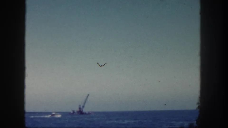 1962 San Diego 8mm frame showing a distant crane barge and small boat wake on the ocean, viewed through dark foreground edges.