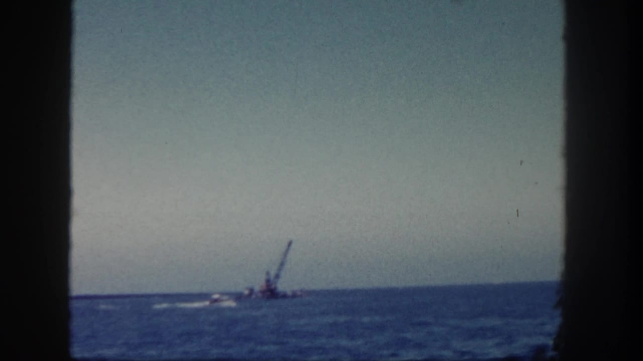 1962 San Diego coastal footage frames a distant crane barge on blue water under a pale sky, with dark vertical edges in view.