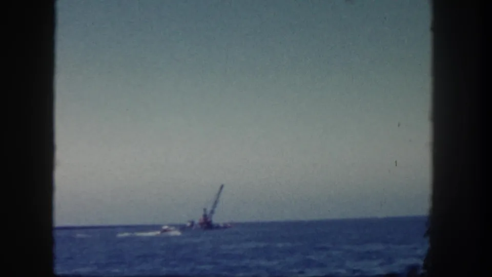 1962 San Diego coastal footage frames a distant crane barge on blue water under a pale sky, with dark vertical edges in view.