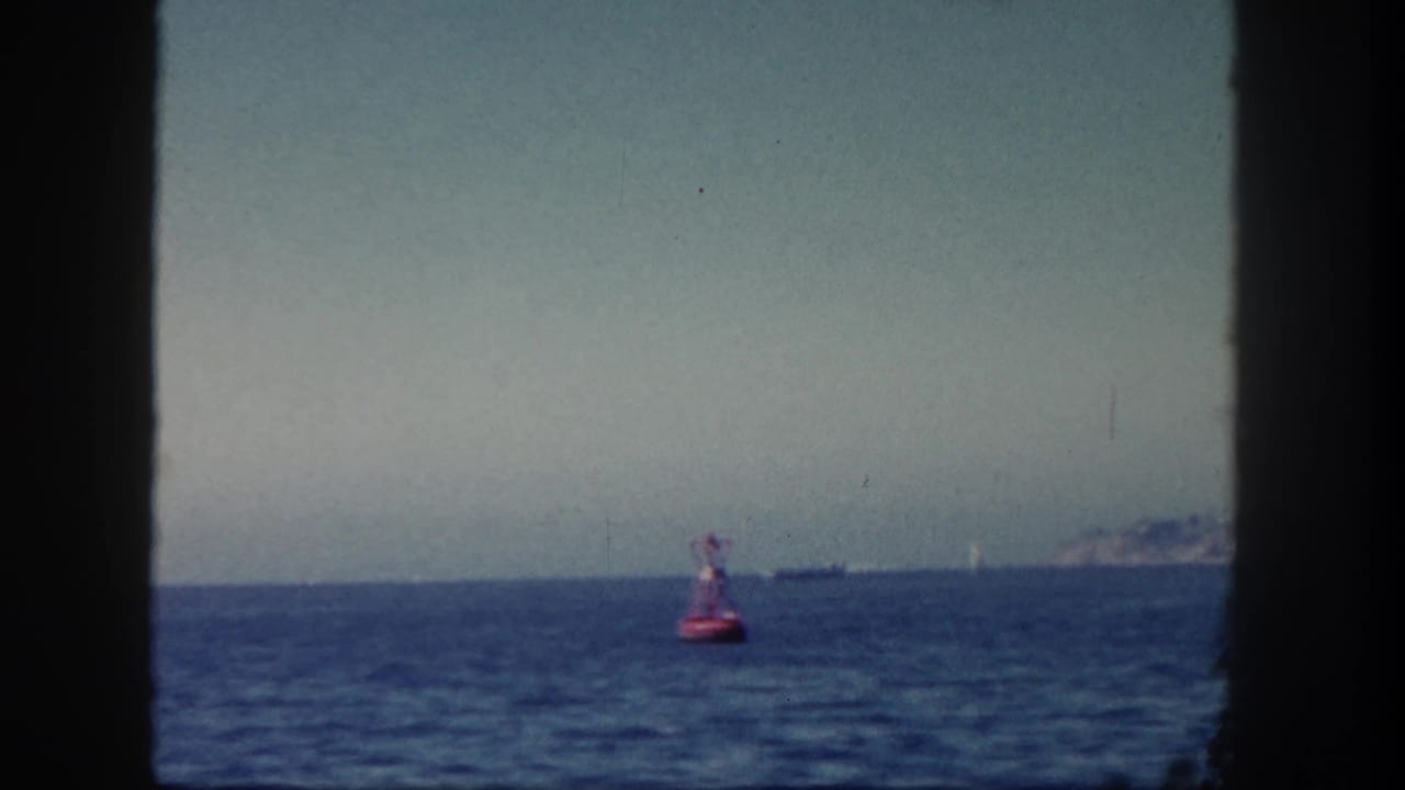 Red buoy floating on blue sea with distant ships and coastline under a hazy sky.