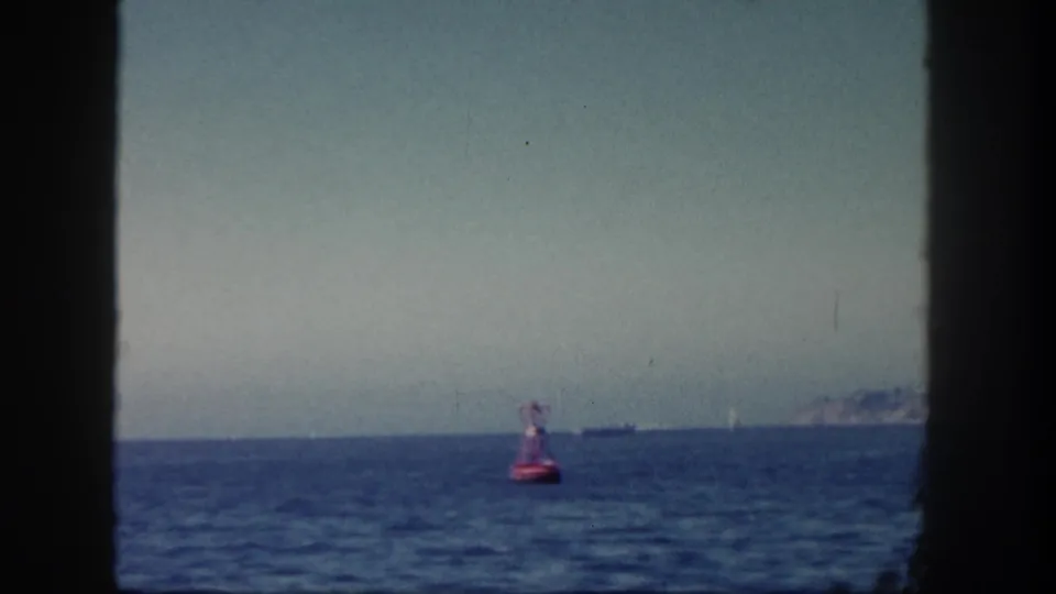1962 San Diego 8mm frame showing a red buoy on open water, distant vessels, hazy sky, and faint coastline through dark framing.