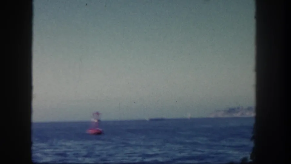 1962 San Diego home-movie view of a red buoy in the ocean, with a distant ship and pale coastline beneath a hazy sky.