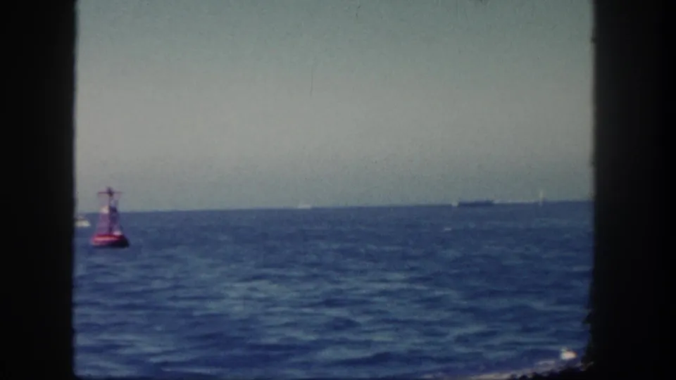 1962 San Diego coastal frame showing a red buoy, blue sea, distant ship, faint sail-like shape, and hazy shoreline through dark framing.