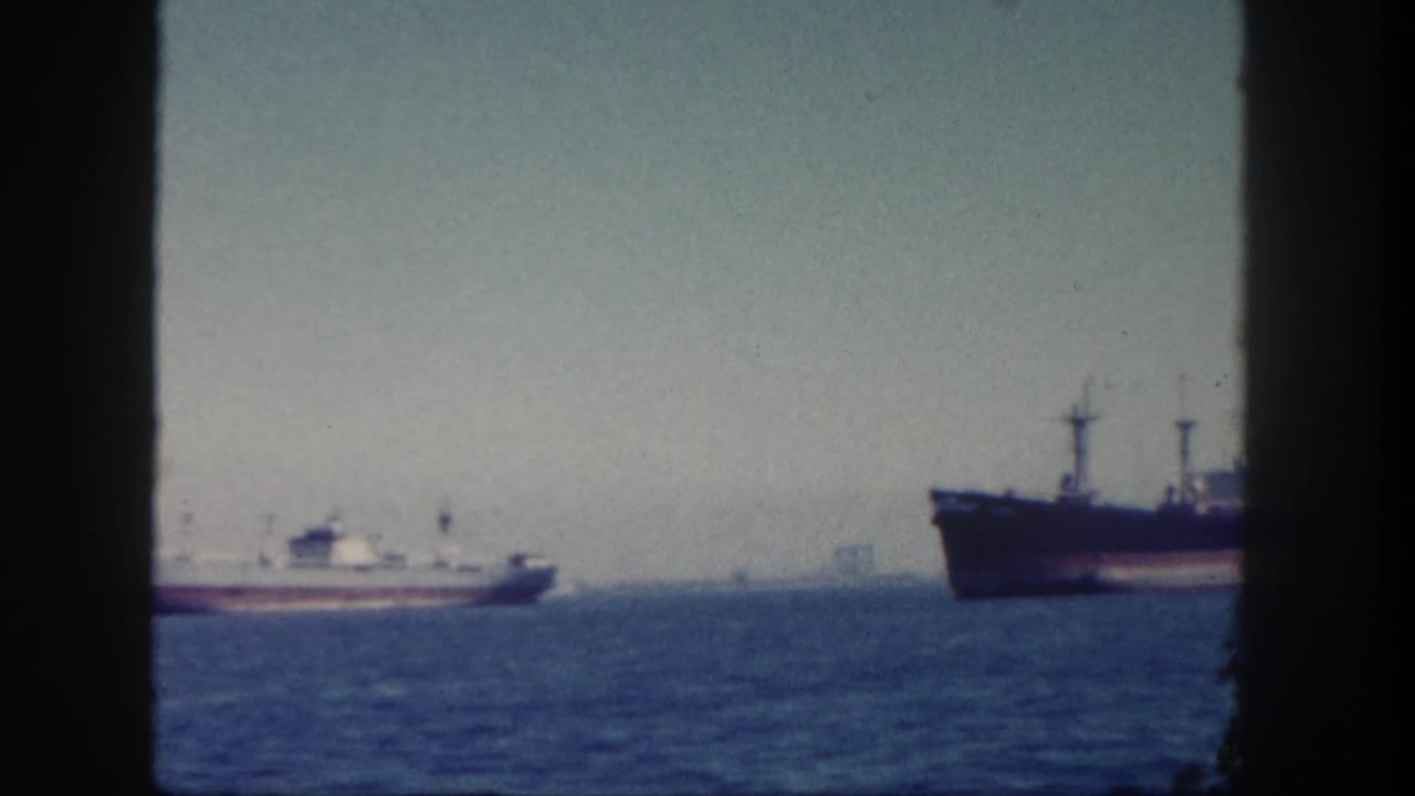 1962 San Diego harbor view with a small white-and-red vessel moving offshore between dark frame edges and a larger ship at right.