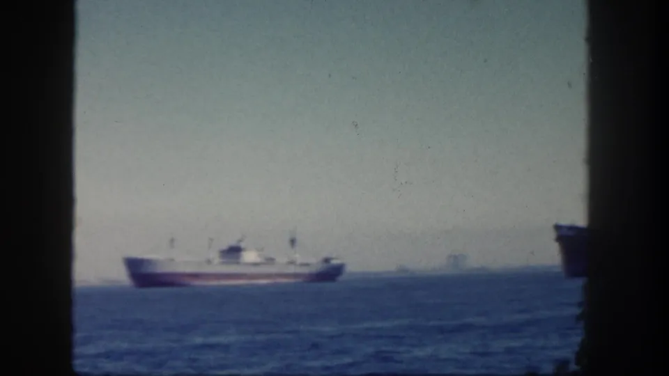 1962 San Diego 8mm still showing a white-and-red vessel offshore, bordered by dark vertical foreground shapes and a hazy skyline.