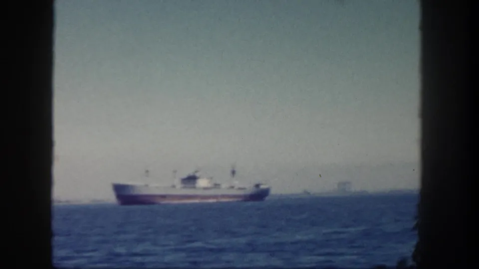 1962 San Diego harbor view of a red-hulled cargo ship offshore, framed by dark window mullions under a hazy sky.