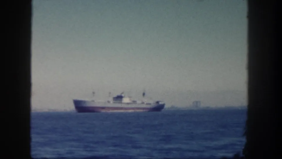 The 8mm frame shows the cargo ship farther across blue water, partly composed against dark window framing.