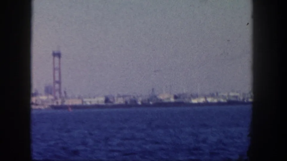 1962 San Diego coastal view from a boat, showing blue water, hazy sky, distant industrial shoreline, and a red bridge or tower structure.