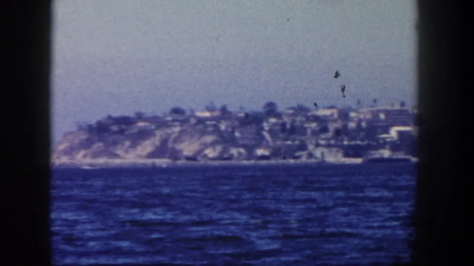 1962 San Diego-area 8mm view from a boat toward a hazy cliffside shoreline with houses above blue water.