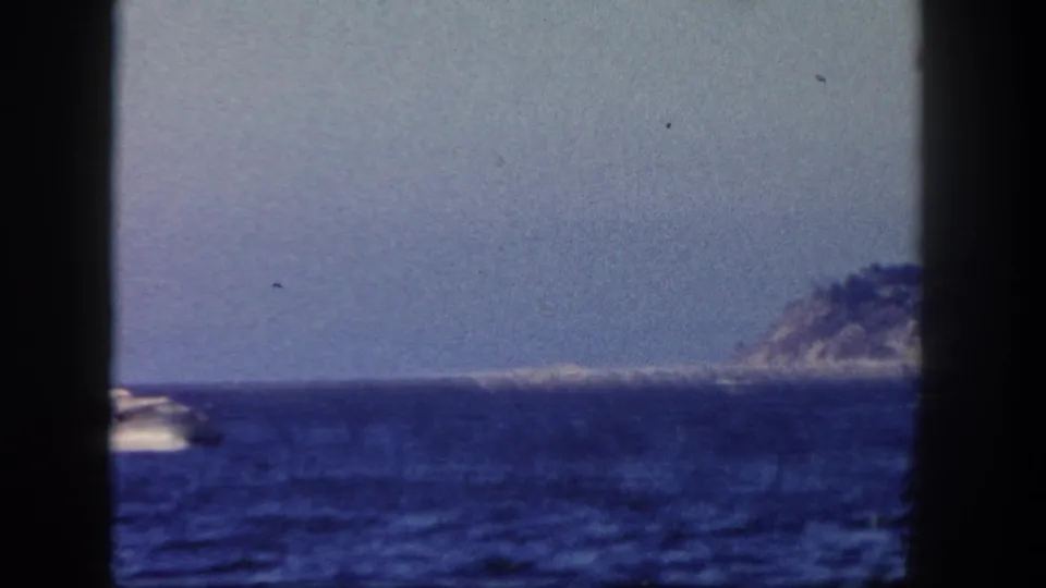 1962 San Diego-area 8mm frame showing a small motorboat crossing blue water with distant shoreline hills beyond dark gate edges.