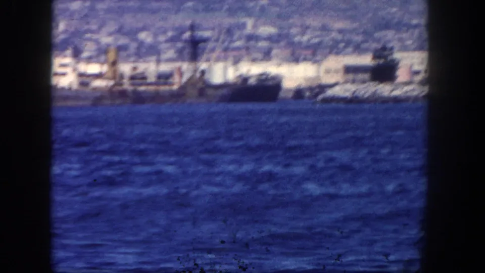1962 San Diego-area harbor frame showing a larger vessel near waterfront structures, viewed across blue water on 8mm film.