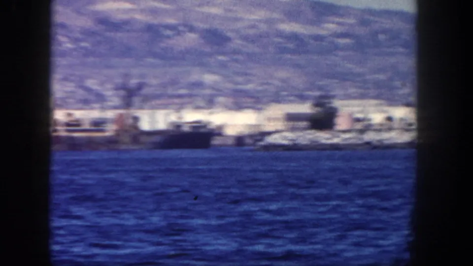 1962 San Diego waterfront seen from the water, with a docked ship, shoreline buildings, blue ocean, and hills in the background.