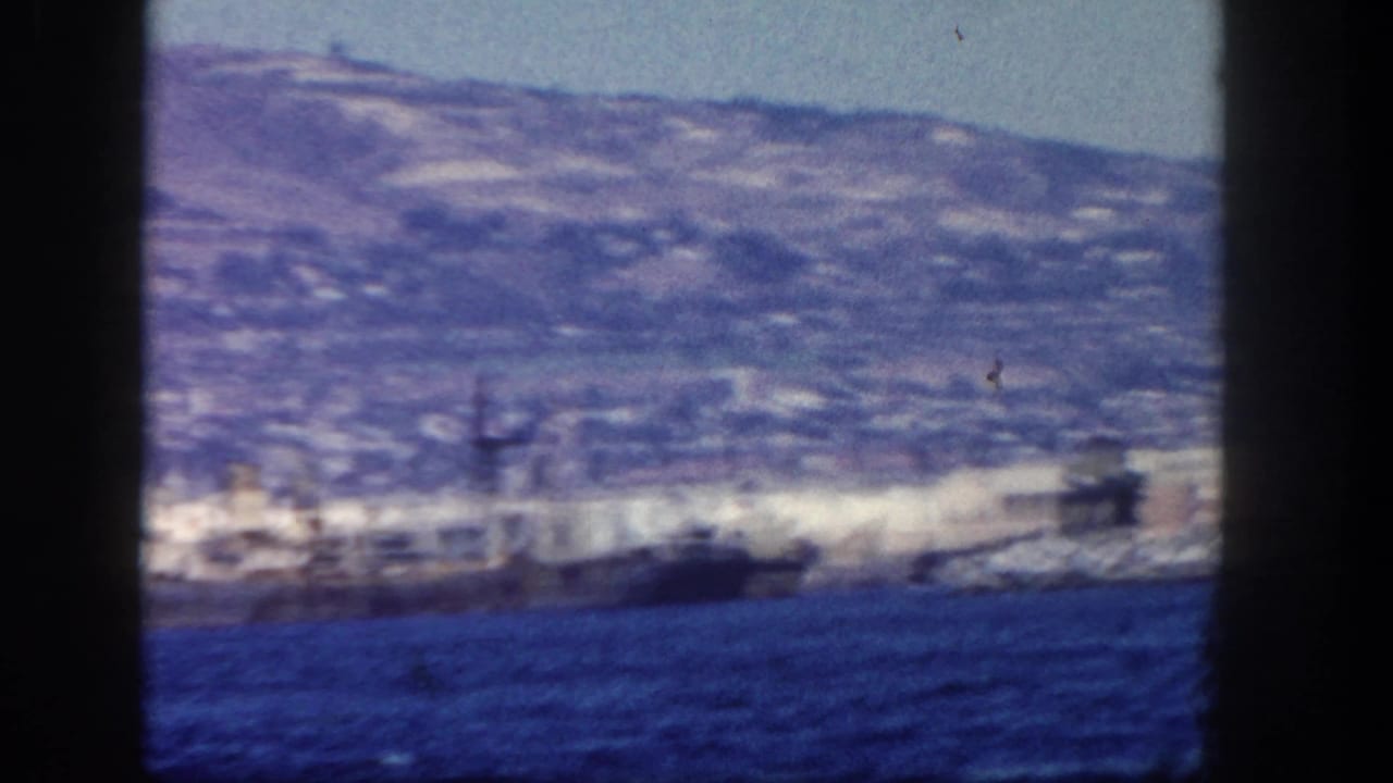 Blurred waterfront buildings and a ship along the shore with hills beyond.