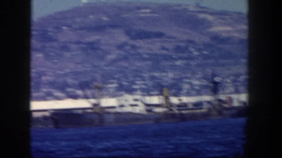 1962 home-movie view across San Diego waterfront, with water in front, low shoreline structures, and a broad hillside background.