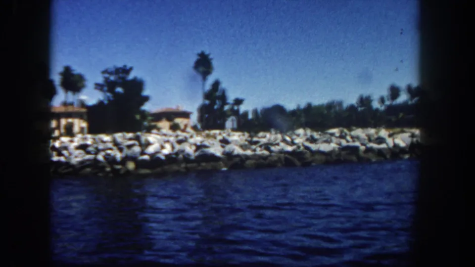 San Diego waterfront scene with a rock breakwater, palm trees, low buildings, dark blue water, and clear sky in a 1962 8mm frame.