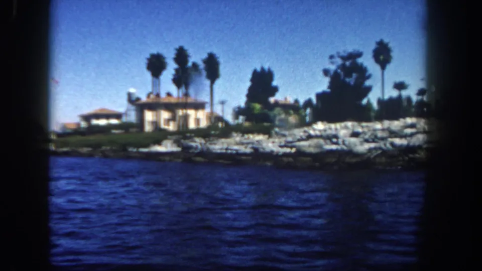 1962 San Diego coastal home-movie frame with blue water, a low waterfront house, palms, rocks, and dark 8mm frame edges.