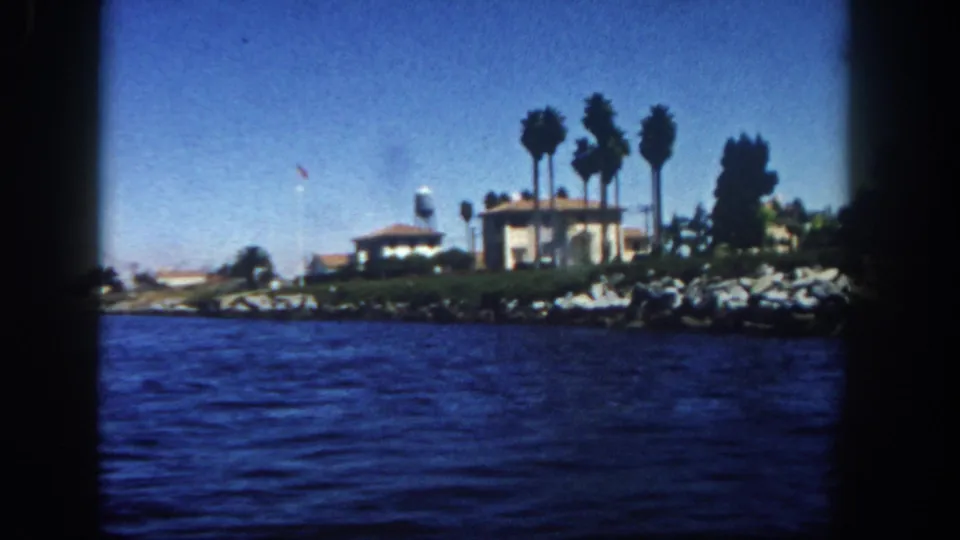 1962 San Diego shoreline from a boat, showing a stucco house, palms, rock-lined edge, water, and a small water tower in the distance.