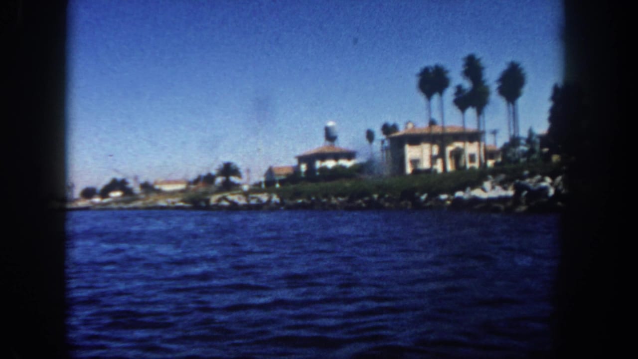 1962 San Diego boat view across blue water toward shoreline homes, tall palms, rocks, and a water tower.
