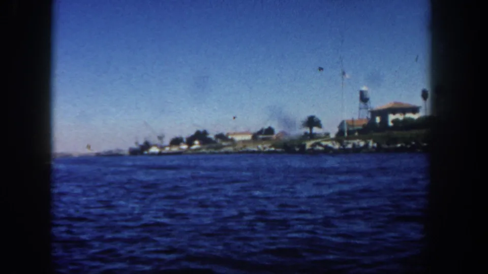 Distant 1962 San Diego shoreline from a moving boat, showing blue water, low waterfront buildings, palms, and a water tower.