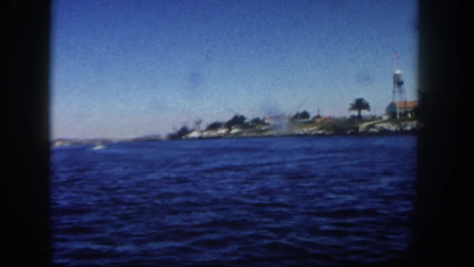 Wide 1962 San Diego boat-tour frame with rippled blue water, low shoreline, palms, buildings, and a water tower.